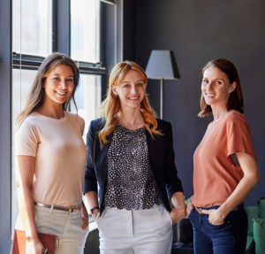 Portrait of happy businesswomen standing by window. Smiling female professionals are in brightly lit creative office. They are wearing smart casuals.